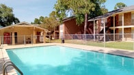 The outdoor pool area serves as a central amenity within the community, featuring a spacious rectangular swimming pool filled with clear, blue-tinted water. A concrete pool deck surrounds the water, providing ample space for lounging and movement. The area is fully enclosed by chain-link fencing, ensuring a secure environment.

Mature trees are positioned throughout the surrounding grounds, casting dappled shade across the deck and nearby lawn areas, contributing to a comfortable outdoor atmosphere. Well-maintained green grass extends beyond the pool enclosure, offering additional open space within the community.

A covered pavilion structure is visible nearby, providing a shaded gathering spot adjacent to the pool. Concrete picnic tables and seating are situated poolside, offering convenient outdoor dining and relaxation options. Entry points with metal handrails provide safe access into the pool.

The surrounding brick residential buildings, featuring two-story construction with covered walkways, balconies, and exterior staircases, frame the pool area on multiple sides. The overall setting benefits from abundant natural light and an open sky, creating a pleasant and functional outdoor recreational space.