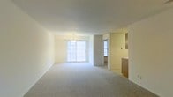 This single-floor space presents an open and airy layout centered around a bright, carpeted living area. The room is finished with neutral cream-toned walls and smooth ceilings, creating a clean and understated aesthetic throughout. A classic chandelier hangs as a subtle focal point, adding a touch of traditional charm to the space.

Natural light flows generously into the living area through a large sliding glass door fitted with grid-patterned glazing, complemented by an additional window nearby that further brightens the interior. The soft, light-toned carpet underfoot contributes to the room's warm and inviting atmosphere.

Toward the side of the living area, a partial wall separates the space from what appears to be an adjacent area, offering a sense of defined zones while maintaining an open feel. A glimpse into the kitchen reveals warm wood-toned cabinetry, hinting at a functional and practical cooking space. The overall layout flows naturally from the living area toward these adjoining spaces, making the floor plan feel connected and well-organized. The space provides a versatile, move-in ready environment with a timeless, neutral design suitable for a variety of interior styles.
