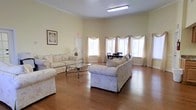This single-floor space offers a comfortable and well-appointed common area designed for relaxation and gathering. The room is anchored by warm hardwood flooring that runs throughout, lending a classic and inviting feel to the interior. Soft, neutral-toned walls in a pale yellow hue create a calm and airy atmosphere, complemented by crown molding that adds a refined architectural touch.

The seating area features multiple floral-upholstered sofas arranged around a wrought-iron and glass coffee table, providing ample space for conversation and lounging. A dining area is visible in the background, furnished with a dark wood table and matching chairs, making the space functional for both social gatherings and meals.

Natural light floods the room through several large windows dressed with layered sheer and draped curtains, softening the light while maintaining brightness throughout the day. Overhead fluorescent lighting ensures the space remains well-lit at all times. A framed artwork piece decorates one wall, adding a personal and homely touch. An exit door is also visible, indicating this space is part of a larger facility. Overall, the space exudes a warm, welcoming ambiance suited for community use.