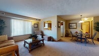 This single-floor residence on Harbour Run S in Mentor-on-the-Lake, Ohio, offers a well-organized layout with distinct living, dining, kitchen, bedroom, and bathroom areas. The living room is generously sized, featuring wall-to-wall carpeting, a textured ceiling, and a large window dressed with vertical blinds that allows natural light to fill the space. A pass-through opening connects the living area to the kitchen, adding an open, communicative feel between rooms. The kitchen is equipped with dark wood cabinetry, ample counter space, a gas range, refrigerator, and a window above the sink. Tile-style flooring, a decorative backsplash, and a compact dining nook complete the kitchen's functional character. The separate dining area accommodates a full dining set beneath a pendant light fixture, with open sightlines toward the kitchen and hallway. The bedroom is comfortably sized with carpeted floors, a large window, and a spacious closet featuring bifold doors, offering generous storage. The full bathroom includes a single-sink vanity with wood cabinetry, a mirrored medicine cabinet, and a tub-shower combination. Throughout the residence, neutral tones and consistent carpeting create a cohesive and comfortable ambiance.