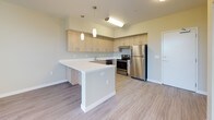 This contemporary single-floor residence is thoughtfully designed with a clean, modern aesthetic and practical layout. The kitchen serves as a central highlight, featuring light wood cabinetry extending to the upper walls for generous storage, crisp white quartz countertops, and a full suite of stainless steel appliances including a refrigerator, range, and dishwasher. Pendant lighting hangs above the kitchen peninsula, which provides additional prep space and doubles as a casual dining area. Light-toned wood-look flooring runs throughout the main living and kitchen areas, lending warmth and a cohesive feel to the open-concept space. The bedroom is carpeted for comfort and benefits from a large window that fills the room with natural light and frames a view of surrounding greenery. The bathroom is finished with a wood-grain vanity, dark quartz countertop, a large mirror, and a full-sized soaking tub with overhead shower. An in-unit laundry closet accommodates a side-by-side washer and dryer, adding everyday convenience. The overall palette of soft neutrals, natural wood tones, and crisp whites creates a calm, inviting atmosphere throughout the residence.