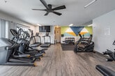 Located at 1311 Washington Pl in Santa Ana, CA, this well-equipped fitness center offers a comprehensive workout environment designed for a variety of exercise routines. The single-floor facility features durable wood-look flooring throughout, creating a clean and modern aesthetic.

The cardio zone is outfitted with multiple treadmills and elliptical machines, allowing several users to train simultaneously. A wall-mounted flat-screen TV provides entertainment during workouts. A ceiling fan overhead helps maintain comfortable airflow throughout the space. Natural light flows in through a large sliding glass door, which opens to a view of a sparkling outdoor pool and surrounding greenery, adding a refreshing outdoor connection to the workout experience.

The strength training area includes a multi-function cable machine and an additional functional trainer, both offering versatile resistance-based exercises. A dedicated free weights section features a full rack of dumbbells alongside an adjustable weight bench, catering to a range of strength training needs. Rubber flooring mats are placed beneath the free weight area for added safety and stability.

A bold geometric mural in teal, yellow, and white anchors one wall, adding an energetic visual focal point. Floor-to-ceiling mirrors amplify the sense of space and allow users to monitor their form. The facility is well-lit with recessed and overhead fluorescent lighting throughout.