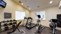 This single-floor fitness room is designed to provide a functional workout space with a focused selection of exercise equipment. The room features light cream-colored walls and recessed ceiling lighting that creates a bright, energizing atmosphere. Natural light filters in through multiple windows fitted with roller shades, contributing to the room's open feel.

The flooring consists of durable, rubberized material well-suited for exercise use. The cardio section is equipped with two treadmills and an elliptical trainer, offering users a variety of cardiovascular workout options. A weight bench and a rack of dumbbells spanning a range of sizes are positioned along one wall, providing strength training capabilities.

Ventilation is well-addressed through multiple ceiling-mounted HVAC vents, ensuring a comfortable environment during workouts. An informational notice is posted on the wall, likely outlining facility rules or equipment usage guidelines. The space is efficiently organized to maximize usability while maintaining clear pathways between equipment, making it practical for individual or small-group use.