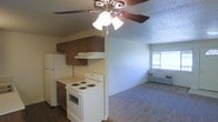 This single-floor space offers a practical and functional layout, combining the kitchen and living area in an open, connected arrangement. The kitchen is equipped with a white electric range, a refrigerator, and a range hood, all complemented by warm, dark wood cabinetry that provides ample storage. The kitchen features easy-to-clean hard flooring, which transitions into wall-to-wall carpeting in the adjoining living area, creating a clear distinction between the two zones. A ceiling fan with an integrated light fixture is centrally positioned overhead, ensuring both air circulation and ambient lighting throughout the shared space. The living area benefits from a window that invites natural light, enhancing the overall brightness of the room. A wall-mounted heating unit is also present, offering convenient climate control. The textured ceiling adds a classic touch to the interior. Overall, the space presents a straightforward, comfortable layout well-suited for everyday living, with all essential features thoughtfully integrated into a compact and efficient single-level floor plan.