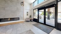Located at 1 Chapel St in Nanaimo, BC, this thoughtfully designed lobby and common area serves as an inviting gateway for residents and visitors alike. The space is organized across a single floor and encompasses a welcoming living room lounge, and two connecting hallways.

The main lounge area immediately impresses with its soaring double-height ceilings and an abundance of natural light streaming through expansive floor-to-ceiling windows. A sleek, linear gas fireplace set against large-format stone tile cladding serves as a focal point, flanked by warm wood accents and complemented by a contemporary pendant light overhead. Comfortable seating — including a grey sofa, modern accent chairs, and oval wood coffee tables — creates an inviting atmosphere, while curated wall art adds a refined, artistic touch throughout.

Light grey large-format porcelain tile flooring runs seamlessly through the lounge and into both hallways, lending the space a clean, cohesive aesthetic. The primary entry hallway features glass double doors that open to the street, maximizing light and visibility. A secondary hallway offers practical amenities including a parcel locker system, recycling stations, elevator access, and bulletin boards, ensuring residents have everything they need conveniently at hand. Overall, the space strikes a balance between modern elegance and everyday functionality.