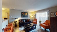 This single-floor residence offers a thoughtfully designed layout that balances comfort and functionality across several distinct living areas. The living room serves as the heart of the home, featuring warm hardwood-style flooring, neutral gray walls, and a sliding glass door with vertical blinds that allows natural light to fill the space and provides access to an outdoor balcony. The kitchen showcases dark navy cabinetry, granite-style countertops, a double stainless steel sink, and a striking linear mosaic tile backsplash that adds a modern, polished touch. Adjacent to the kitchen, a dedicated dining area offers an intimate setting with hardwood-style flooring, framed wall art, and a decorative mirror that enhances the sense of space. The bedroom is a serene retreat, finished with soft carpeting, a large window dressed with elegant curtains that invite natural light, and neutral tones that create a calming ambiance. The bathroom features a full tub and shower combination with clean white tile surround. Completing the unit, an in-unit laundry area includes a top-loading washer and built-in shelving above for additional storage. Throughout, the space maintains a cohesive, contemporary aesthetic with warm accent lighting and thoughtful design details.
