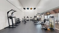 Located at 110 Mountainhigh Dr in Nashville, TN, this well-appointed fitness center offers a comprehensive workout environment within a generously sized, single-room layout. The space is defined by impressively high ceilings that create an open, airy atmosphere, keeping the area feeling expansive and comfortable during exercise. The crisp white walls are complemented by dark rubber flooring, providing both durability and safety underfoot. Recessed ceiling lights, along with decorative acoustic panels in teal and gold, add a modern, polished touch to the interior design. A large mirror wall along one side of the room visually doubles the space while offering views into an adjacent amenity area with vaulted, wood-beamed ceilings. Equipment is thoughtfully arranged throughout, including multiple treadmills, an elliptical trainer, a stationary bike, a cable machine, selectorized strength machines, a multi-function pull-up and dip tower, adjustable benches, and a tiered dumbbell rack with a full range of free weights. A wall-mounted flat-screen television enhances the experience for cardio users, while posted workout charts provide additional guidance. The overall ambiance is clean, bright, and purpose-built for fitness.