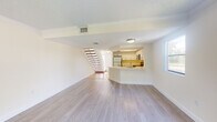 This two-story space is designed with a clean, open layout that promotes a bright and airy atmosphere throughout. The main level centers around an open-concept living and kitchen area, where light wood-style flooring flows seamlessly underfoot. The kitchen is well-appointed with white shaker cabinetry, light stone countertops, stainless steel appliances including a range, microwave, and refrigerator, and a peninsula that separates it from the living space. A tray ceiling detail adds architectural interest above the kitchen. The living area extends toward a sliding glass door that opens to a private balcony, offering outdoor views and abundant natural light. Crown molding accents the living space, adding a refined touch. A staircase leads to the upper floor, where multiple generously sized rooms feature the same light wood-style flooring, large windows with vertical blinds, and textured ceilings. Several rooms include ceiling fans, and views of lush greenery are visible from the windows. Throughout both floors, neutral warm-toned walls and consistent flooring create a cohesive, move-in-ready environment well-suited for comfortable everyday living.