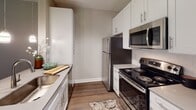 Located at 6500 Grand Retreat Dr. in Roanoke, Virginia, this single-floor residence offers a thoughtfully designed layout with a clean, contemporary aesthetic throughout. The kitchen is a functional highlight, featuring stainless steel appliances including a refrigerator, electric range, and over-the-range microwave, paired with crisp white cabinetry, gray countertops, and warm wood-look flooring that extends into the adjoining living areas. The open transition from the kitchen to the main living space creates a generous, versatile area ideal for everyday use and entertaining. Neutral warm-beige walls and white trim provide a cohesive palette throughout the unit. The bedrooms are carpeted and bathed in natural light through large windows — several of which frame lush, wooded views, offering a serene natural backdrop. The bathroom is well-appointed with a white vanity, gray countertop, a tub-shower combination, and matching wood-look flooring. Additional closet storage and a hallway connecting the key living zones contribute to the unit's practical and organized flow. Overall, the space presents a balanced combination of modern finishes and comfortable livability in a residential community setting.
