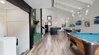 This two-floor space is designed with entertainment and recreation at its core, combining striking architectural details with thoughtfully curated amenities. The main level is anchored by a dramatic vaulted ceiling with exposed white-painted beams, flooded with natural light through large windows and a glass door. Wood-look plank flooring runs throughout, complementing the crisp white vertical shiplap walls accented by bold black trim — a design palette punctuated by deep green brick, creating a distinctive lodge-inspired aesthetic.

The main entertainment area features a full-size pool table as a centerpiece, alongside a bar-height table and a wall-mounted TV. A dark-toned kitchen island with bar seating and a fully equipped kitchen with stainless steel refrigerator and black range provide functional hosting capabilities. The dining area accommodates multiple guests with glass-top tables and rich emerald velvet chairs beneath a sputnik chandelier.

A dedicated gaming room houses arcade machines and large-screen TVs with racing-style gaming chairs, while a lounge area offers plush dark velvet sofas on a bold teal patterned rug. A staircase leads to the upper mezzanine level, overlooking the main floor through black-railed balcony, adding vertical drama to the overall space.