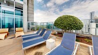 Located at 848 Spring St NW in Atlanta's Midtown neighborhood, this rooftop amenity level offers an impressive outdoor retreat with sweeping views of the Atlanta skyline, including notable landmarks such as Georgia Tech's campus. At the heart of the space is a generously sized swimming pool with crystal-clear turquoise water, framed by a broad concrete pool deck lined with blue lounge chairs ideal for sunbathing. The surrounding architecture features a striking combination of exposed concrete, brick, and large floor-to-ceiling glass panels, lending the space a modern, industrial aesthetic. A covered pergola area with warm wood detailing and string lights provides a shaded lounge zone furnished with contemporary tables and chairs set on artificial turf, perfect for casual gatherings. Manicured topiaries and landscaped greenery add natural accents throughout the deck. Glass railings along the perimeter ensure unobstructed panoramic views of the cityscape on all sides. The space is well-suited for both relaxation and socializing, offering a dynamic urban outdoor environment elevated above the city.