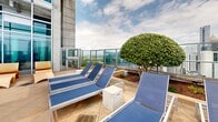 Located at 848 Spring St NW in Atlanta's Midtown neighborhood, this rooftop amenity level offers an impressive outdoor retreat with sweeping views of the Atlanta skyline, including notable landmarks such as Georgia Tech's campus. At the heart of the space is a generously sized swimming pool with crystal-clear turquoise water, framed by a broad concrete pool deck lined with blue lounge chairs ideal for sunbathing. The surrounding architecture features a striking combination of exposed concrete, brick, and large floor-to-ceiling glass panels, lending the space a modern, industrial aesthetic. A covered pergola area with warm wood detailing and string lights provides a shaded lounge zone furnished with contemporary tables and chairs set on artificial turf, perfect for casual gatherings. Manicured topiaries and landscaped greenery add natural accents throughout the deck. Glass railings along the perimeter ensure unobstructed panoramic views of the cityscape on all sides. The space is well-suited for both relaxation and socializing, offering a dynamic urban outdoor environment elevated above the city.