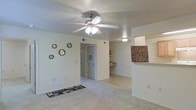 This single-story space offers a straightforward, functional layout centered around a carpeted living area. The main living space is highlighted by a ceiling fan with an integrated light fixture, providing both air circulation and ambient lighting. Neutral-toned walls and light-colored carpet lend a clean, understated aesthetic throughout.

A defining architectural feature is the pass-through opening on the right side of the living area, which connects to the kitchen and provides a sense of openness between the two spaces. Built-in shelving is visible in the transitional area, offering convenient display or storage options. The kitchen counter is partially visible through the pass-through, suggesting a practical and accessible layout.

A doorway at the far end of the living area leads to additional rooms, providing a clear sense of flow through the space. Vents are positioned in the ceiling for efficient climate control. Décor accents, including circular wall mirrors and decorative signage, add a personal touch to the otherwise neutral interior. The overall ambiance is modest, bright, and livable, suited for everyday comfort.