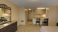 This single-floor unit offers a practical and efficient layout designed to maximize the use of available space. The open-concept living area features neutral-toned carpet flooring and warm beige walls, creating a cohesive and understated aesthetic throughout. A textured ceiling adds subtle visual interest overhead, while built-in wall vents provide climate control throughout the space.

The kitchen is positioned along one wall and is equipped with light-colored cabinetry offering ample storage, a sink, and counter space. A peninsula-style breakfast bar with seating for two defines the boundary between the kitchen and the living area, making it a convenient spot for casual dining or workspace use. Under-cabinet and overhead fluorescent lighting ensure the kitchen remains well-lit and functional.

A hallway extends from the main living area, flanked by closet doors that provide generous storage options. The hallway leads to the rear of the unit, where additional rooms can be accessed. The overall ambiance of the space is clean, straightforward, and move-in ready, with a neutral color palette that lends itself well to a variety of furnishing styles. The layout is well-suited for those seeking a low-maintenance, efficiently organized living environment.