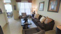 The living area of this single-floor space is thoughtfully designed to create a warm and inviting atmosphere. At its center, a plush dark charcoal sofa is complemented by a matching oversized armchair, both arranged around a sleek dark wood coffee table that anchors the room. The seating is dressed with an assortment of textured throw pillows in neutral and earthy tones, adding depth and comfort to the arrangement.

A large window dressed with elegant rust-toned curtains and vertical blinds serves as the focal point of the room, flooding the space with natural light and creating an airy feel. The soft, light-colored carpeting underfoot enhances the room's warmth and comfort.

Decorative elements are carefully curated throughout the space. A decorative mirror in a dark frame is mounted on the wall, reflecting light and adding visual depth. A stylish table lamp with a dark sculptural base provides ambient lighting, while a centerpiece floral arrangement on the coffee table introduces a natural, organic touch.

The overall ambiance is relaxed yet refined, with a cohesive neutral color palette punctuated by warm accent tones, making this living space both functional and aesthetically appealing.