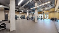 Located at 808 Constance St in New Orleans, Louisiana, this single-floor fitness facility offers a generously sized, open-plan layout designed to accommodate a comprehensive range of workout activities. The space is defined by its industrial-inspired aesthetic, featuring soaring ceilings with exposed ductwork and HVAC systems, structural concrete columns, and sleek linear pendant lighting that illuminates the floor throughout.

The facility is organized into distinct functional zones. A dedicated cardio area is positioned along a wall of large, multi-pane windows, flooding the space with natural light and providing views of the surrounding cityscape. This section houses a row of treadmills, elliptical trainers, and other cardiovascular equipment. Adjacent to this, a strength training zone features a comprehensive selection of cable machines, weight racks, barbells, kettlebells, medicine balls, and selectorized resistance equipment. Full-length mirrors line the walls in multiple areas, enhancing the sense of openness and supporting proper form during workouts.

The flooring throughout consists of durable rubber gym material, suitable for heavy use. A water fountain station and multiple access doors are conveniently integrated along one wall. Accent walls in warm yellow tones add visual energy to the space, complementing the overall modern, utilitarian design of this well-equipped fitness center.
