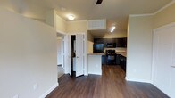 This single-story space offers a functional and cohesive open-concept layout, where the main living area flows seamlessly into the kitchen. The flooring throughout is a rich, dark hardwood-style material that adds warmth and contrast to the neutral, light-colored walls. Crown molding details lend a polished architectural touch, while a ceiling fan in the living area provides both practicality and a subtle design accent.

The kitchen is well-appointed with dark espresso-toned cabinetry that extends to the upper walls, providing ample storage. Black appliances, including a refrigerator, range, and over-the-range microwave, are neatly integrated into the layout. Light-toned countertops contrast the dark cabinetry, and a peninsula-style counter serves as a natural divider between the kitchen and the living space. Track lighting above the kitchen ensures the area is well-lit.

Adjacent to the main living area, a set of bifold doors opens to a laundry closet, where a washer and dryer are conveniently housed. A hallway leads to additional rooms, including a carpeted bedroom visible in the background, offering a comfortable contrast to the hard flooring in the main living spaces.