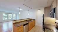 Located at 87 New St in Cambridge, MA, this well-appointed two-bedroom, two-bathroom residence offers a thoughtfully designed single-level layout. The open-concept kitchen is a standout feature, boasting rich granite countertops, warm honey-toned cabinetry, tile flooring, and a full suite of stainless steel appliances including a refrigerator, dishwasher, electric range, and microwave. Dual pendant lights hang above the counter, adding a refined touch to the space. The kitchen flows naturally toward the living area, which features beautiful hardwood floors that reflect natural light streaming through a wide set of windows overlooking lush, mature greenery — a serene and private outlook. Both bedrooms offer plush carpeting and ample natural light; the primary bedroom includes a tall wooden wardrobe and multiple windows, while the second bedroom benefits from the same warm, light-filled atmosphere. The two bathrooms are finished in clean, neutral tones with tile flooring; one includes a full tub-shower combination with a curved curtain rod. A connecting hallway ties the layout together efficiently, making the overall flow of the residence both practical and comfortable.