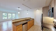 Located at 87 New St in Cambridge, MA, this well-appointed two-bedroom, two-bathroom residence offers a thoughtfully designed single-level layout. The open-concept kitchen is a standout feature, boasting rich granite countertops, warm honey-toned cabinetry, tile flooring, and a full suite of stainless steel appliances including a refrigerator, dishwasher, electric range, and microwave. Dual pendant lights hang above the counter, adding a refined touch to the space. The kitchen flows naturally toward the living area, which features beautiful hardwood floors that reflect natural light streaming through a wide set of windows overlooking lush, mature greenery — a serene and private outlook. Both bedrooms offer plush carpeting and ample natural light; the primary bedroom includes a tall wooden wardrobe and multiple windows, while the second bedroom benefits from the same warm, light-filled atmosphere. The two bathrooms are finished in clean, neutral tones with tile flooring; one includes a full tub-shower combination with a curved curtain rod. A connecting hallway ties the layout together efficiently, making the overall flow of the residence both practical and comfortable.