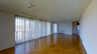 This single-floor space presents a clean, open layout characterized by its warm parquet hardwood flooring arranged in a classic block pattern, which adds texture and visual interest underfoot. The most prominent architectural feature is the expansive wall of floor-to-ceiling windows fitted with vertical blinds, spanning nearly the full length of the main living area. These oversized windows allow an abundance of natural light to fill the room, creating a bright and airy atmosphere throughout the day. A secondary window opening is positioned along the adjacent wall, further enhancing ventilation and light distribution. The ceilings are smooth and relatively generous in height, contributing to the spacious feel of the interior. The walls are finished in a neutral white tone, providing a versatile backdrop that complements the warm hues of the flooring. A baseboard heating unit is visible beneath the windows, indicating a practical heating solution integrated discreetly into the design. The overall layout is open and unobstructed, lending itself well to flexible furniture arrangements. The space maintains a straightforward, functional aesthetic with a focus on light and openness.