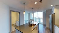 This two-bedroom, two-bathroom residence offers a thoughtfully designed single-floor layout with a seamless blend of modern aesthetics and functional living. The open-concept kitchen serves as a central hub, featuring a large island with a sleek quartz countertop and a deep undermount sink finished in brushed gold. Pendant lights with glass shades hang above the island, adding a refined touch to the space. White cabinetry and contemporary hardware complete the kitchen's clean, modern look, while wood-look flooring flows throughout the unit.

The living area is bathed in natural light through expansive floor-to-ceiling windows that frame sweeping urban skyline views, creating a bright and airy atmosphere. The generous glazing provides a strong visual connection to the cityscape beyond.

A bathroom is conveniently accessible from the main living area, finished with white cabinetry, a dark countertop, and a full mirror. The overall ambiance of the unit is modern and polished, with neutral tones, recessed lighting, and thoughtful design details that create a cohesive, contemporary living environment.