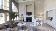 Located at 100 Crescent Arbor Dr in Cary, NC, this single-floor space offers a refined and welcoming atmosphere defined by thoughtful design and upscale finishes. The main living area immediately draws attention with its dramatically high ceilings, which amplify the sense of openness and grandeur throughout the room. A striking floor-to-ceiling fireplace serves as the focal point, framed by a classic white mantel and topped with a built-in niche that elegantly houses a mounted television. The warm, neutral tones of the walls complement the soft, sophisticated furnishings, creating an inviting and cohesive aesthetic. Large arched windows along one side of the room flood the interior with generous natural light, offering views of lush greenery outside and seamlessly connecting the indoors with the surrounding landscape. The space is thoughtfully arranged to accommodate both relaxed seating and a more intimate dining area, making it versatile for a variety of uses. Decorative accents, including table lamps, framed artwork, and carefully selected furnishings in muted patterns, contribute to the space's polished yet comfortable character. The overall ambiance is one of understated elegance, balancing architectural distinction with a warm and livable environment.