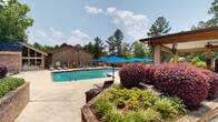 Located at 1811 Sibley Rd in Augusta, Georgia, this outdoor amenity area is centered around a generously sized swimming pool framed by well-maintained landscaping, including ornamental shrubs, colorful flowering plants, and mature trees that provide a natural backdrop. The pool deck offers ample space for sunbathing, lined with cushioned lounge chairs positioned to take in the open sky and surrounding greenery.

Adjacent to the pool is a large covered pavilion that serves as an outdoor gathering and entertainment hub. The pavilion features a white beadboard ceiling outfitted with multiple ceiling fans, ensuring comfort during warm Georgia summers. Ceiling-mounted televisions provide entertainment throughout the space. Seating options are varied and plentiful, ranging from high-top bar tables and chairs to Adirondack-style seating arrangements set atop decorative area rugs, creating distinct social zones within the pavilion.

A standout feature of the pavilion is a brick-faced outdoor fireplace, complete with a rustic wood mantel and decorative accents, adding warmth and ambiance for cooler evenings. The brick architectural detailing is carried throughout the surrounding structures, including the adjacent clubhouse buildings, which feature dramatic angled rooflines and arched detailing. The entire space is enclosed by brick columns and fencing, with manicured hedges contributing to a refined, resort-like atmosphere.