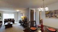 This single-floor residence offers a welcoming and functional layout centered around an open-concept living and dining area. The dining space is highlighted by an elegant chandelier with multiple light fixtures that cast a warm glow over the dark wood dining table and matching lattice-back chairs, creating an inviting atmosphere for meals and gatherings. A console table adorned with a classic table lamp and decorative accents lines the wall, adding character and warmth to the space.

The living area flows seamlessly from the dining room, featuring plush carpeting in a neutral tone that extends throughout the main living spaces, contributing to a cozy, cohesive feel. Natural light filters in through a window, brightening the room and complementing the soft, neutral wall palette. A textured ceiling adds subtle architectural detail throughout.

A warm-toned wooden interior door provides access to additional areas of the home, with a hallway visible beyond leading to further rooms. The overall design balances practicality with comfort, utilizing neutral tones, warm lighting, and classic furnishings to create a livable and welcoming environment. The open layout between the primary living spaces promotes easy flow and makes the home feel spacious and connected.