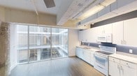 This single-floor loft-style space showcases a compelling blend of industrial and contemporary design. The open-concept layout is immediately striking, anchored by a expansive floor-to-ceiling glass wall that floods the interior with natural light and offers views into a multi-story atrium below, creating a unique sense of connectivity and openness.

The exposed ceiling is a standout architectural feature, revealing raw beams, structural pipes, and ductwork finished in white, lending the space an authentic industrial character. The light hardwood flooring runs throughout, providing a warm contrast to the cool, neutral palette of the walls and ceiling.

The kitchen is thoughtfully integrated along one wall, featuring white cabinetry, a double-basin sink, a white electric range, and a built-in microwave. The clean, minimalist cabinetry and gray countertops maintain a cohesive, modern aesthetic throughout.

The open living area benefits enormously from the atrium-facing glass wall, with diffused natural light creating a bright and airy ambiance. The overall atmosphere is modern and urban, with the industrial ceiling details and glass architecture working together to deliver a visually dynamic and functional space.