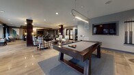 Located at 360 Creamery Way in Exton, Pennsylvania, this single-floor amenity lounge is a thoughtfully designed social and recreational space that blends contemporary style with a welcoming atmosphere. The interior is anchored by polished large-format tile flooring throughout, recessed lighting, and a neutral gray color palette accented by warm wood tones and curated artwork.

The game room area serves as a central focal point, featuring a full-size billiards table beneath a sleek pendant light, a dedicated poker or card table with upholstered seating, and a classic arcade game station set against a dark stone accent wall. A wall-mounted flat-screen TV and framed sports-themed artwork complete the entertainment atmosphere.

Adjacent to the game area, a comfortable lounge zone offers plaid and patterned armchairs arranged for conversation, complemented by a rustic wood-slatted ceiling detail and a large-scale abstract painting. A media console with a mounted television adds to the relaxed ambiance.

A separate dining or meeting area provides a dark wood table with woven rattan chairs, bathed in natural light from floor-to-ceiling windows dressed in sheer navy curtains with outdoor views of the surrounding community. Architectural columns with dark wood cladding punctuate the open layout, adding structure and visual interest throughout the space.