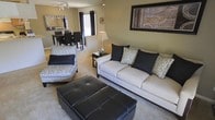 This single-story space showcases an open-concept floor plan that seamlessly connects the living and dining areas, creating a cohesive and functional layout. The living area is anchored by a cream upholstered sofa adorned with black and patterned accent pillows, complemented by a matching accent chair and a tufted black leather ottoman that serves as a central coffee table. Neutral beige carpeting runs throughout, contributing to a warm and inviting atmosphere. A framed piece of wall art adds a decorative focal point above the sofa, while track lighting on the ceiling provides targeted illumination. The dining area, visible from the living space, features a dark-toned dining table and chairs, with a window allowing natural light to filter in. Adjacent to the dining space, a glimpse of the kitchen reveals additional cabinetry and countertop space, suggesting a practical and well-connected layout. The consistent neutral color palette of warm beige walls and dark accent furnishings throughout creates a balanced, contemporary aesthetic that feels both polished and livable.