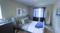 This single-floor residence offers a comfortable and thoughtfully designed living space. The bedroom serves as a highlight of the home, featuring warm neutral-toned walls that create a calm and inviting atmosphere. A large window dressed with bold dark curtains allows natural light to filter into the room while providing privacy, and the contrast between the drapes and the soft wall color adds a modern, sophisticated touch. The room comfortably accommodates a full-sized bed dressed in crisp white bedding accented with blue and yellow throw pillows, adding pops of color to the serene palette. Flanking the bed are stylish tripod floor lamps that provide ambient lighting and contribute to the room's contemporary aesthetic. Two framed art prints featuring urban architectural motifs hang side by side above the headboard, reinforcing the modern design sensibility. Carpeted flooring adds warmth and comfort underfoot. The overall ambiance of this space is relaxed yet polished, blending functional furnishings with decorative details that make it feel welcoming and well-curated — an ideal retreat for rest and relaxation.