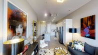 This 1-bedroom, 1-bathroom residence is designed with a modern, open-concept layout that seamlessly connects the living, dining, and kitchen areas into one cohesive space. The interior is finished with smooth, light-toned flooring that runs throughout, contributing to a clean and airy atmosphere. Track lighting along the ceiling provides adjustable, focused illumination across the main living area, complementing the recessed lighting overhead.

The kitchen is a standout feature, outfitted with crisp white cabinetry, a central island, and stainless steel appliances including a refrigerator, microwave, and range. The high-gloss finishes on the cabinetry add a polished, contemporary feel to the culinary space.

The living area is generously proportioned, offering ample room for a full seating arrangement. Bold decorative elements — including a large-scale art piece, gold-accented shelving, and a graphic area rug — lend the space a curated, design-forward character. The ceilings are notably elevated, enhancing the sense of openness and volume throughout.

A corridor leads to the private bedroom and bathroom, providing a clear separation between living and sleeping areas. Overall, the unit presents a sophisticated, well-appointed interior that balances modern aesthetics with functional living.