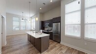 This single-floor residence showcases a contemporary open-concept design that seamlessly connects the kitchen and living areas. The kitchen is a standout feature, centered around a large island with a white quartz countertop, an undermount sink, and dark espresso cabinetry. Stainless steel appliances — including a French door refrigerator, electric range with a subway tile backsplash, and built-in microwave — complete the space. Pendant lighting hangs above the island, while recessed lighting and a ceiling fan illuminate the broader living area. Tall windows throughout invite abundant natural light, enhancing the airy feel created by the notably high, vaulted ceilings.

Wood-look plank flooring runs throughout the main living and kitchen spaces, transitioning to plush carpet in the bedrooms. The bedrooms feature ceiling fans, generous wall space, and access to walk-in closets with built-in shelving and rod systems. The bathroom is finished with dark cabinetry, quartz countertops, a large mirror, and wood-look tile flooring. A hallway connects the private sleeping quarters to the main living area, providing a well-organized and functional layout throughout.