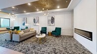 This expansive single-floor amenity space is thoughtfully designed to serve as a sophisticated communal hub, blending comfort, functionality, and contemporary style throughout. The main lounge area is anchored by a coffered ceiling with a striking gold-finished tray detail, elegant wainscoting, and a sleek linear fireplace flanked by open shelving — all contributing to an upscale, welcoming atmosphere. Rich teal accents appear throughout, from accent chairs and cabinetry to herringbone-patterned tile backsplashes, creating a cohesive and vibrant design language. The social kitchen features a large quartz-topped island with a bold green base, warm wood cabinetry, a built-in media display, and pendant lighting — ideal for gatherings and casual socializing. A dedicated dining area offers a refined setting with botanical-print wallpaper, a deep teal tray ceiling, a sculptural brass chandelier, and floor-to-ceiling windows overlooking lush outdoor greenery. Private study rooms enclosed in steel-framed glass doors provide quiet, focused work environments. Wide, art-lined corridors connect each zone, displaying curated abstract works on clean white walls over light wood flooring. The mail lobby offers a welcoming reception area with a white desk and full-wall mailboxes beneath a green accent ceiling. Throughout, light hardwood flooring and recessed lighting unify the space with a clean, modern finish.