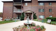 This well-maintained courtyard serves as a charming and inviting outdoor space set against the backdrop of a multi-story brick building. At its centerpiece stands an elegant tiered stone fountain, surrounded by colorful seasonal plantings including vibrant red blooms and ornamental shrubs that add warmth and visual interest to the landscape. Neatly trimmed evergreen trees flank a covered entrance with a classic columned portico, creating a polished and welcoming focal point. The walkways are paved and well-defined, providing easy accessibility throughout the space. On the periphery, green Adirondack chairs are thoughtfully placed beneath the canopy of a large, mature shade tree, offering a comfortable spot to relax and enjoy the outdoors. Additional seating areas can be found along the building's edge, further enhancing the space's usability. The courtyard benefits from open sightlines to the surrounding neighborhood, with mature trees lining the adjacent streets and providing a sense of natural enclosure. Upper-level balconies overlook the courtyard, integrating the outdoor and indoor environments seamlessly. Overall, the space strikes a balance between formal landscaping and casual comfort, making it an appealing communal outdoor area.