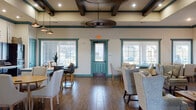 Located at 5215 S Raul Longoria Rd in Edinburg, Texas, this single-story community clubhouse is thoughtfully designed to serve as a hub for socializing, recreation, and productivity. The grand entry foyer sets an inviting tone with soaring ceilings, a decorative tiled floor inset, rich wood-look flooring, teal-accented trim and doors, and arched passageways that lead deeper into the facility.

The main lounge area features coffered ceilings with teal-painted beams, a ceiling fan, and abundant natural light streaming through multiple windows with horizontal blinds. A comfortable seating arrangement surrounds a barn-door-style TV console, creating a relaxed gathering atmosphere. Adjacent to the lounge, a dining and social area provides bar-height and standard seating for residents to gather informally.

A dedicated game room offers round tables and upholstered chairs beneath patterned carpet, decorated with framed playing card artwork, and includes a wall-mounted TV and a board game library for resident entertainment. The computer lab is equipped with multiple workstations, teal ergonomic chairs, and patterned carpet, offering practical tech access.

A multipurpose room with light flooring, folding tables, and a whiteboard easel supports group activities, classes, and meetings, and connects to a small kitchenette. Throughout the space, the consistent teal and warm neutral palette creates a cohesive, welcoming environment.