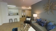 This single-floor space showcases an open-concept design that effortlessly connects the living area and kitchen, creating a cohesive and functional layout. The living area is carpeted and thoughtfully arranged, featuring a neutral-toned sofa accented with blue throw pillows, a sleek leather accent chair, and a contemporary glass and chrome coffee table. A large floor mirror adds depth to the space, enhancing the sense of openness. Track lighting on the ceiling provides flexible illumination throughout, complemented by warm ambient lighting from a stylish table lamp. The accent wall, painted in a deep gray tone, adds a modern contrast to the lighter surrounding walls and ceiling. The kitchen is partially visible from the living area, separated by a breakfast bar with industrial-style metal stools. White cabinetry and light countertops give the kitchen a clean, bright aesthetic. The overall ambiance of the space is contemporary and polished, with a carefully curated blend of metallic accents, neutral tones, and bold pops of color that contribute to a sophisticated yet comfortable atmosphere.