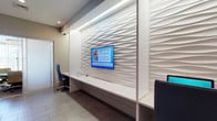 This single-floor amenity space is thoughtfully designed to serve residents with a polished, contemporary aesthetic throughout. The main lobby area sets the tone with large-format tile flooring in neutral tones, recessed lighting, and bold colorful artwork that adds energy to the clean, modern palette. A built-in workstation area features a sleek white countertop desk and an eye-catching textured wave-panel accent wall with integrated cove lighting, creating a striking focal point. A flat-screen display mounted on this feature wall keeps residents informed of upcoming community events. Adjacent to the main area, a dedicated conference or meeting room offers a refined, upscale atmosphere with a round glass-top table, contemporary upholstered chairs, sheer floor-to-ceiling drapery, and abundant natural light streaming through large windows that frame lush outdoor greenery. An elegant chandelier overhead completes the sophisticated setting. The mail room corridor is equally well-appointed, featuring dual rows of stainless steel individual mailboxes lining both walls, vibrant teal upper walls accented with artwork and decorative vases, and tile flooring that continues the cohesive design language. Together, these spaces create a functional yet stylish environment that reflects a high standard of residential community living.
