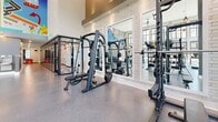 Located at 553 W 100 S in Salt Lake City, Utah, this fitness center is a single-floor exercise facility offering a generous, open layout designed to accommodate a wide range of workout needs. The space is defined by its dramatically high ceilings and floor-to-ceiling grid-pane windows that line the perimeter, flooding the interior with abundant natural light and providing open views of the surrounding urban streetscape.

The flooring throughout is a durable, speckled rubber surface that transitions to a turf section, ideal for functional training and dynamic movement exercises. The main workout area is comprehensively equipped with cardio machines including treadmills, ellipticals, and stationary bikes, alongside a full complement of strength training equipment such as cable machines, Smith machines, power racks, and adjustable benches. A well-stocked dumbbell rack and various free weights round out the offerings.

Ceiling fans and recessed lighting provide both comfort and illumination throughout the space. A striking large-scale, colorful mural depicting mountain landscapes and Utah-inspired imagery serves as a bold focal point along one wall, adding artistic character to the environment. Structural columns punctuate the open floor plan, while mirrored walls in select areas enhance the sense of depth and space. The overall ambiance is modern, energetic, and welcoming.