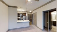 Located at 1570 Atria Ct in Raleigh, NC, this single-floor residence offers a well-designed open-concept layout that seamlessly connects the kitchen and living areas. The kitchen is a standout feature, equipped with stainless steel appliances including a refrigerator, microwave, and range. Dark espresso cabinetry provides a rich contrast against the bright white countertops, while a peninsula with an integrated sink and pendant lighting overhead creates an ideal space for both meal prep and casual dining. A decorative wine rack adds a touch of character to the kitchen backdrop. The living area is finished with plush neutral carpeting, crown molding, and a ceiling fan, contributing to a comfortable and polished atmosphere. Multiple doors visible from the main living space suggest well-distributed storage and private rooms, including a closet with louvered doors. The overall color palette of soft beiges and grays lends the home a clean, cohesive aesthetic. Crown molding throughout adds an architectural detail that elevates the interior finish. The layout is thoughtfully organized to maximize both functionality and livability within a single-story footprint.