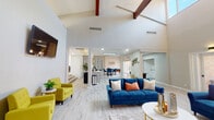 This single-floor space makes a striking architectural statement with its soaring vaulted ceilings accented by bold exposed wood beams. The open-concept layout flows generously from one area to the next, unified by light wood-toned flooring throughout. The main living area is anchored by a dramatic floor-to-ceiling brick fireplace wall, adding warmth and texture, while plush blue velvet sofas and vibrant yellow accent chairs create an inviting, modern atmosphere. A wall-mounted flat-screen television and recessed lighting complete the lounge setting.

The space transitions seamlessly into a dining and game area, where a round pedestal table with deep blue upholstered chairs sits alongside a full-size pool table beneath a ceiling fan. Frosted glass panel partitions and arched architectural details add refined character to this zone. A dedicated wet bar area features white cabinetry with black hardware, a marble-look countertop, a built-in beverage refrigerator, and a coffee station — ideal for entertaining. Natural light filters in through large windows, enhancing the bright, airy ambiance throughout the entire space.