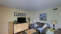 Located at 3975 I-55 N in Jackson, Mississippi, this single-floor space offers a comfortable and inviting atmosphere with thoughtfully arranged interiors. The living area serves as the heart of the space, featuring a plush gray sofa accented with decorative pillows and a soft throw blanket, creating a relaxed and cozy setting. A natural wood dresser with ample drawer storage anchors the entertainment area, housing a flat-screen television alongside lush greenery and decorative dried botanicals that add organic texture to the room. A woven coffee table sits at the center of the space, complementing the earthy tones throughout. Wall art adds personality and color to the neutral palette — a trio of black-and-white framed prints lines one wall, while a vibrant blue floral canvas brings a pop of color to another. A soft table lamp provides warm ambient lighting, enhancing the comfortable feel of the room. The light-toned walls and smooth ceiling keep the space feeling open and airy. Overall, the space strikes a balance between casual comfort and curated style, making it a welcoming environment suitable for relaxing or entertaining.
