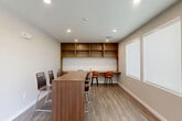 Located at 12121 Greenspoint Drive in Houston, Texas, this single-room space offers a clean, contemporary design well-suited for dining, collaboration, or casual work. The room is finished with warm wood-look luxury vinyl plank flooring that contrasts nicely against the neutral, light-toned walls and crisp white trim, creating a bright and inviting atmosphere.

A central high-top communal table with bar-height chairs serves as the focal point, providing ample seating for group gatherings or shared meals. Along one wall, a continuous built-in wooden counter runs the full width of the room and is accompanied by a set of open-cubby shelves mounted above, offering both functional workspace and display storage. A pair of orange upholstered bar stools anchor this counter area, adding a pop of color to the otherwise neutral palette.

Large windows fitted with roller shades line the adjacent wall, allowing for adjustable natural light throughout the day. Recessed ceiling lighting supplements the natural light, ensuring the space remains well-lit at all times. A glass-panel door provides access to adjacent areas, enhancing the sense of connectivity. The overall ambiance is modern, clean, and functional — ideal for communal dining or a collaborative work setting.