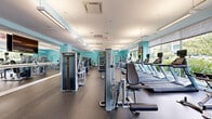 Located at 1 Glover Ave in Norwalk, Connecticut, this expansive fitness center offers a generously sized, single-floor layout designed to accommodate a full range of workout needs. The facility is finished with signature aqua-toned walls that lend a bright, energizing ambiance throughout the space, complemented by vibrant, colorful geometric artwork that adds a modern aesthetic touch.

The fitness floor is comprehensively equipped with a wide array of Matrix-branded cardio machines, including treadmills, ellipticals, stationary bikes, and rowing machines, arranged to maximize both usability and traffic flow. A dedicated strength training zone features multiple selectorized resistance machines and free weight stations, supported by large wall-mounted mirrors that enhance the sense of openness and allow for proper form monitoring.

Durable rubber flooring covers the entire workout area, providing both safety and comfort. Ample natural light pours in through expansive floor-to-ceiling windows along multiple walls, offering pleasant outdoor views of surrounding greenery and the neighboring streetscape. Overhead, a combination of pendant and recessed fluorescent lighting ensures the space is well-lit at all times.

A glass-door entry leads to an adjacent room that appears to serve as a secondary amenity or relaxation area. The overall design is clean, functional, and modern, making this fitness center a well-appointed facility for comprehensive daily training.