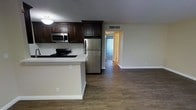 This single-floor space showcases an open-concept layout that seamlessly connects the kitchen and living area, creating a functional and cohesive environment. The kitchen is well-appointed with dark espresso-toned cabinetry that extends to the ceiling, offering generous storage capacity. Stainless steel appliances, including a refrigerator, built-in microwave, and gas range, lend a modern, polished aesthetic to the cooking space. A peninsula counter serves as a natural divider between the kitchen and the adjoining living area, providing additional workspace and potential seating. The warm hardwood-style flooring flows continuously throughout the space, adding visual continuity and a sense of warmth. Neutral-toned walls complement the darker cabinetry and create a balanced, inviting atmosphere. A hallway visible from the main living area leads toward additional rooms, with natural light filtering in through what appears to be a sliding glass door or large window at the far end, brightening the space and offering a connection to the outdoors. Ceiling vents suggest a central HVAC system for year-round comfort.