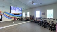 This fitness studio is thoughtfully designed to accommodate a wide range of exercise activities. The space features durable rubberized flooring ideal for high-impact workouts, and the walls are lined with floor-to-ceiling mirrors that enhance the sense of openness while allowing users to monitor their form during exercise. Natural light filters in through multiple windows, creating a bright and energizing atmosphere complemented by recessed and pendant lighting overhead. A large flat-screen display is mounted on the primary wall, enabling users to follow along with workout videos or fitness classes. The studio is well-stocked with equipment, including several stationary cycling bikes for cardio training, a tiered rack of dumbbells in varying weights, and multiple medicine balls organized on dedicated storage racks. A step platform provides additional versatility for aerobic and strength training exercises. The layout is open and spacious, allowing for free movement and group exercise sessions. Informational boards are posted on the walls, offering guidance and resources for users. The overall ambiance is clean, functional, and motivating, making this fitness studio a practical and inviting space for achieving health and wellness goals.