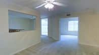 This single-story space offers an open and airy layout, characterized by neutral-toned carpet flooring and light-colored walls that create a bright, welcoming atmosphere. Crown molding runs along the upper perimeter of the main living area, adding a subtle architectural detail that elevates the overall finish of the space. A ceiling fan with a built-in light fixture is centrally positioned overhead, providing both ventilation and ambient lighting.

The living area flows naturally into an adjoining room, separated by a partial wall with a pass-through opening, fitted with track lighting above — likely serving as a functional divide between the living and dining or kitchen areas. This design maintains an open feel while defining distinct zones within the space.

Large sliding glass doors in the main area lead to an outdoor area, allowing generous natural light to pour in and offering views of lush greenery beyond. A second adjacent room mirrors this brightness with its own wide window, also framing outdoor foliage. The consistent use of neutral finishes, crown molding, and natural light throughout gives the space a cohesive, clean, and comfortable character.