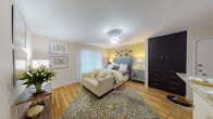 This single-floor studio is thoughtfully laid out to maximize functionality and style throughout its open living space. Warm hardwood flooring runs continuously throughout, lending a cohesive and inviting feel to every area.

The sleeping area serves as the focal point of the space, highlighted by a bold yellow accent wall adorned with decorative floral wall art. A tufted blue upholstered headboard anchors the room, flanked by mirrored nightstands and complementary table lamps. A plush, patterned area rug in grey and yellow grounds the space, while a sliding glass door with vertical blinds allows natural light to fill the room.

Transitioning seamlessly from the bedroom area, the living space offers room for a settee and decorative console table, framed wall art, and a breakfast bar with counter seating — ideal for casual dining or working from home.

The kitchen features rich dark espresso cabinetry, light granite-style countertops, a double basin stainless steel sink, and black appliances including an electric range with overhead ventilation. A pass-through opening connects the kitchen to the main living area, enhancing the open feel.

The bathroom is clean and well-maintained, offering a full tub and shower combination with white tile surround, a toilet, and hardwood-style flooring that carries through from the main living areas. A dark wardrobe cabinet provides additional storage near the sleeping area, rounding out this efficiently designed studio.