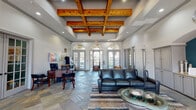 This single-floor community space makes a strong architectural statement from the moment you enter. The main lounge area commands attention with soaring ceilings adorned by a striking coffered grid of warm, natural wood beams, complemented by recessed lighting that highlights the craftsmanship throughout. Expansive arched transom windows line the front facade, flooding the interior with abundant natural light and offering views to the exterior grounds.

The flooring throughout is a rich slate tile laid in a diagonal pattern, with an ornate mosaic inlay near the entryway adding an elegant focal point. The reception and workspace area is positioned near the front, featuring a traditional wood desk and seating, framed by glass-paned French doors that connect seamlessly to adjacent rooms.

A comfortable lounge seating area anchors the main space, furnished with a full leather sofa, accent chairs in teal, and a glass-top coffee table atop layered area rugs. Opposite the seating area, a built-in entertainment wall features floor-to-ceiling gray cabinetry with granite countertops, flanking a wall-mounted television set beneath a decorative arched teal accent wall.

A bright, windowed dining nook with a round table and upholstered chairs provides a more intimate gathering space, bathed in natural light from additional arched French doors. The overall ambiance is polished, welcoming, and designed for both functional and social use.